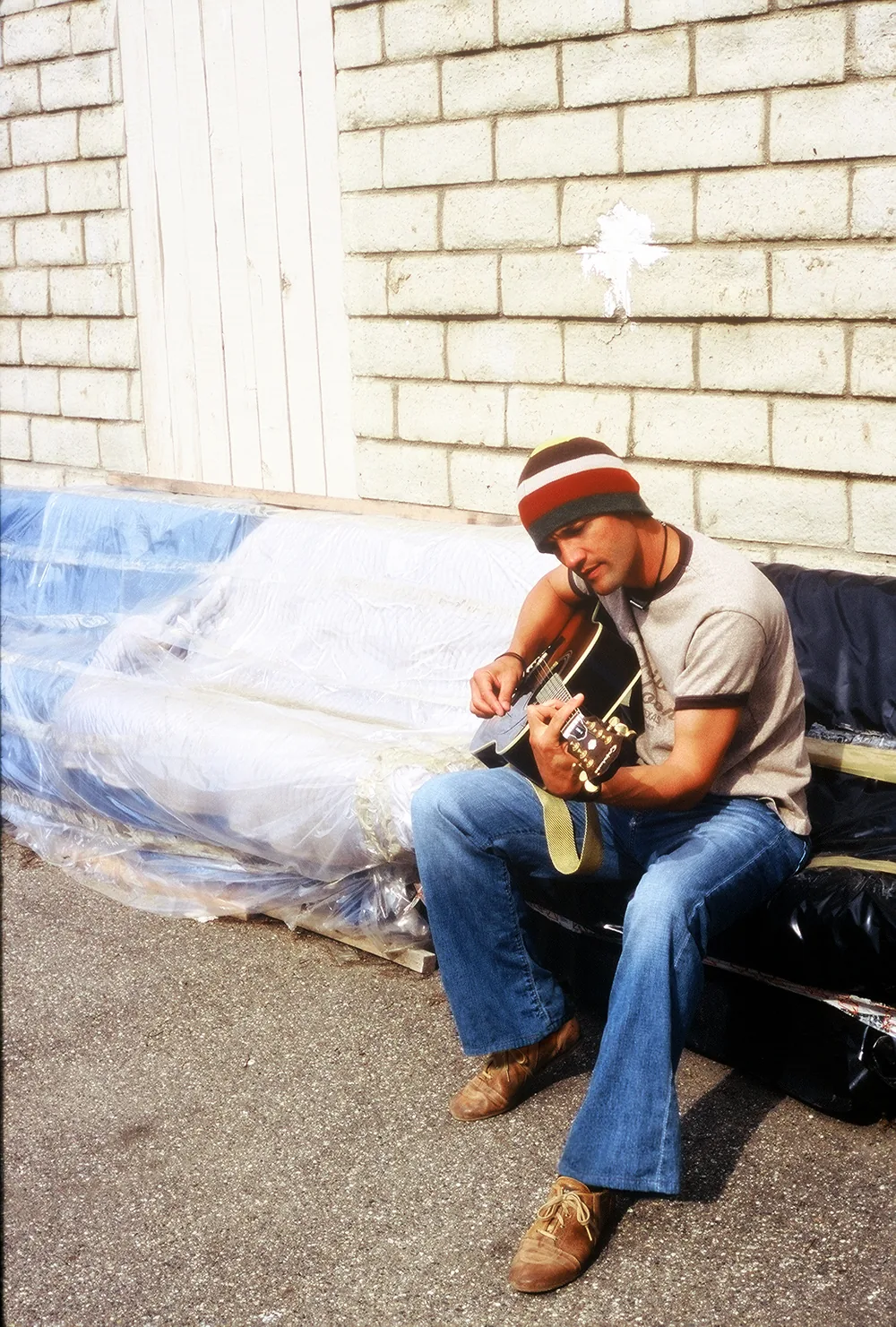 Kef in a striped beanie playing acoustic guitar on a plastic-wrapped couch